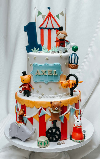 A decorated two-tier circus-themed birthday cake with fondant figures, a red-and-white striped tent topper, a blue number 1, and the name AXEL on a blue plaque, set against a white draped backdrop.